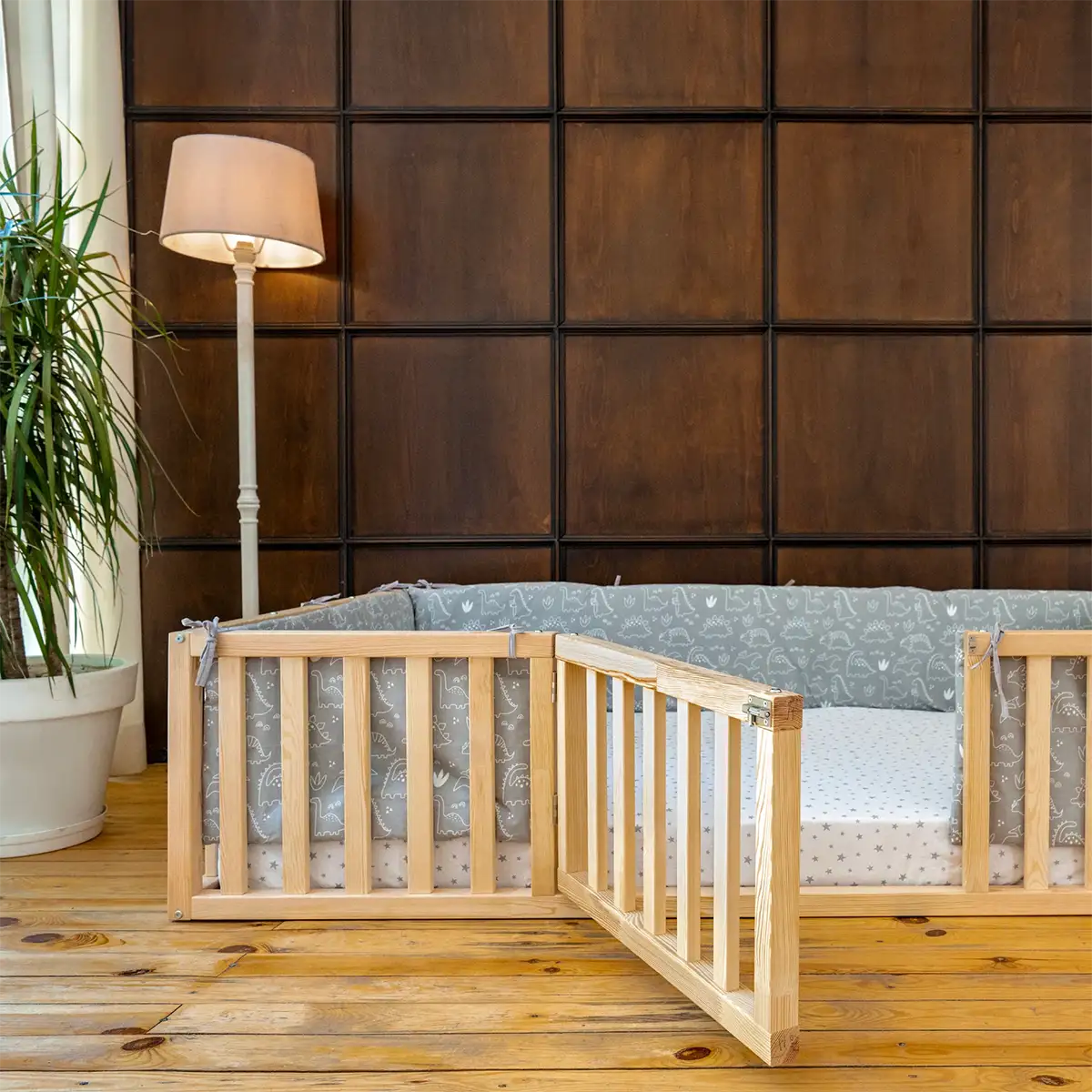 Wooden toddler bed with gray bedding in a room with a brown paneled wall and a lamp.